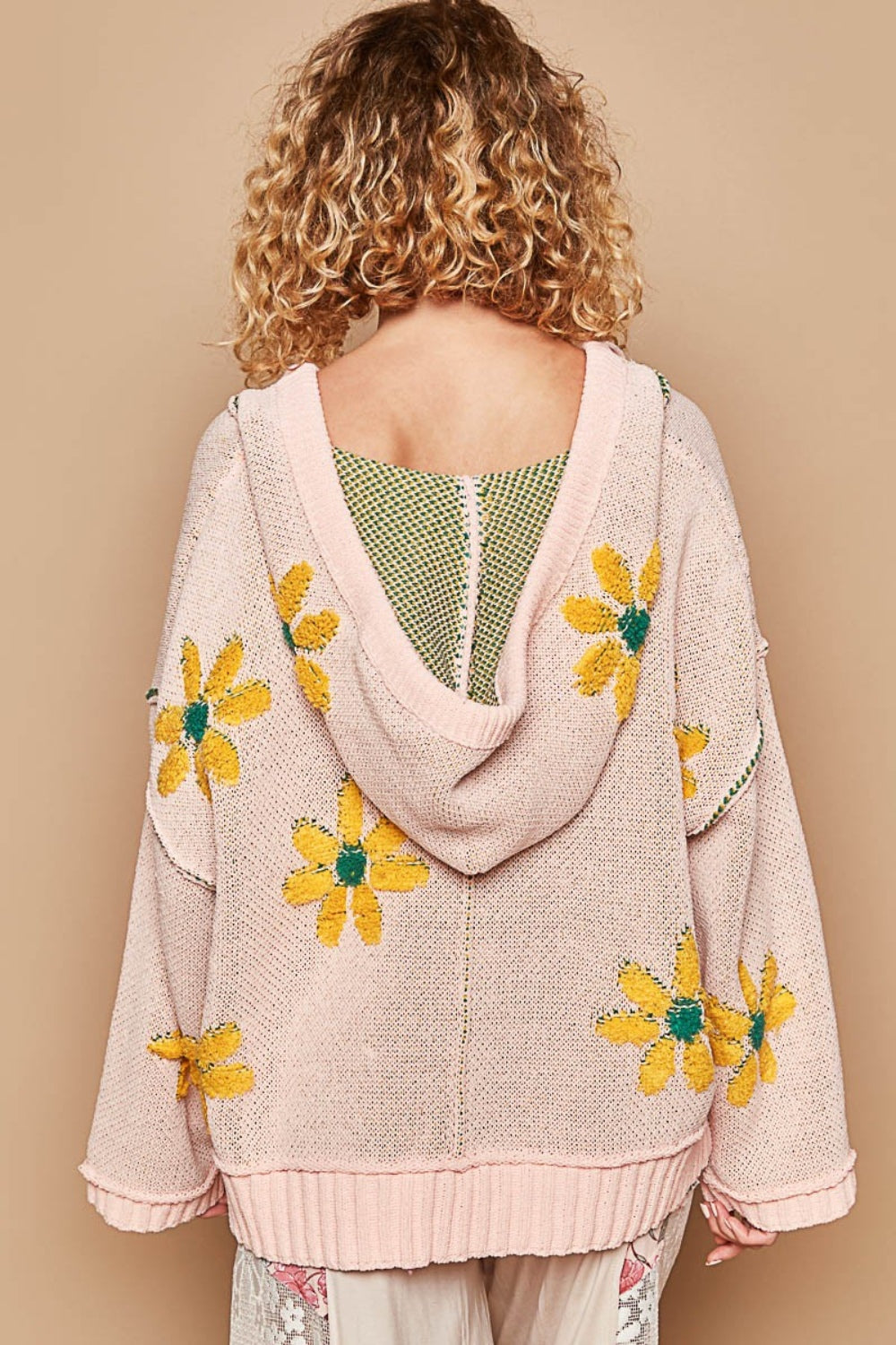 A person with curly blonde hair wears a stylish POL Floral Pattern Hooded High-Low Sweater featuring yellow flower designs and light-colored floral pants. The background is light beige.