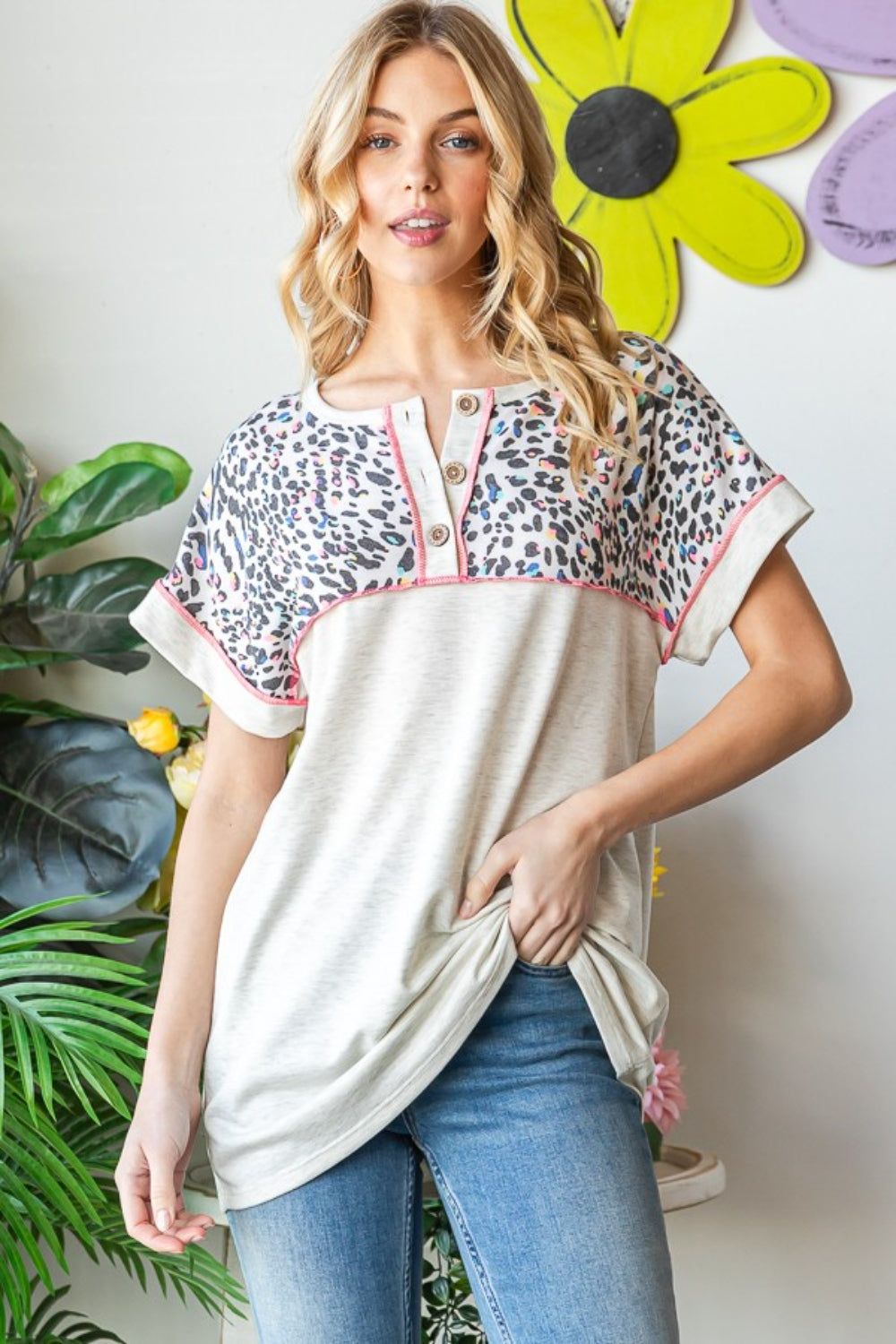 A woman stands indoors wearing a Heimish Full Size Exposed Seam Leopard Short Sleeve T-Shirt with blue jeans, holding the hem of her shirt. She is surrounded by green plants and a wall adorned with yellow and purple flower decorations.