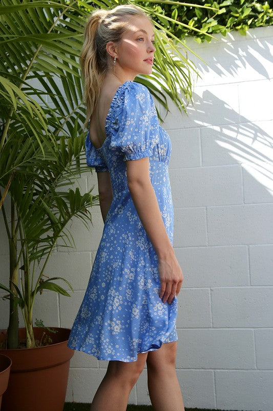 A person wearing the SS puff sleeve dress in a blue floral print stands in front of green palm leaves and a white wall.