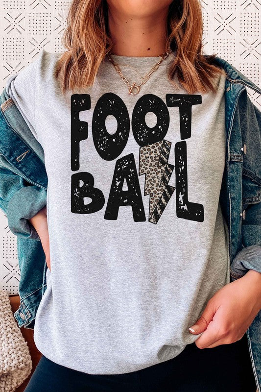 A person wearing a LEOPARD LIGHTNING FOOTBALL Graphic Tee, which features the text "FOOT BALL" and a leopard lightning football design in the center, is standing in a room with a guitar in the background.