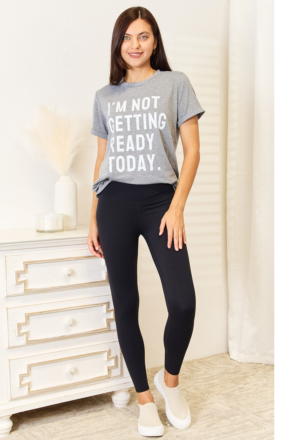 Dressed in a Simply Love I'M NOT GETTING READY TODAY Graphic T-Shirt and vibrant red pants, the individual exudes a carefree style while standing near a white surface adorned with decorative objects.