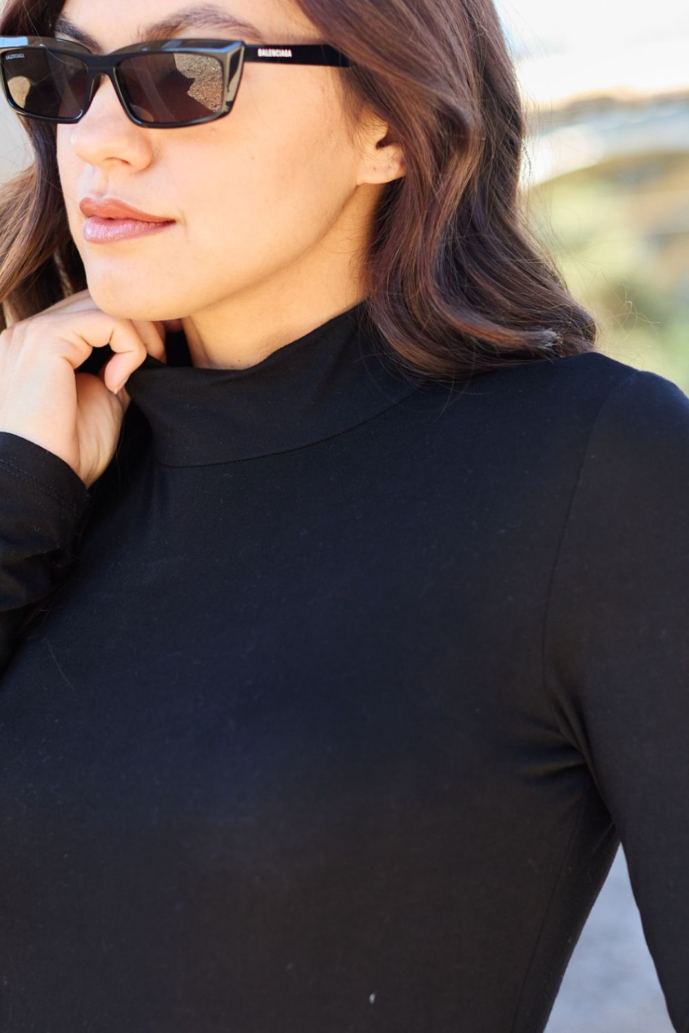 A woman with long, wavy hair stands in front of a light-colored curtain, wearing the Basic Bae Full Size Mock Neck Long Sleeve Bodysuit that offers a basic style and moderate stretch.