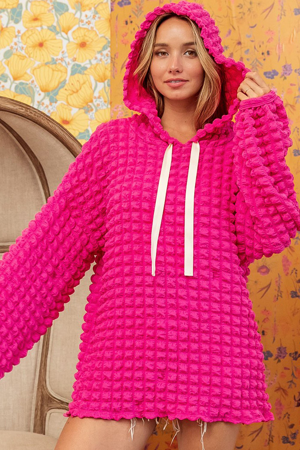 A person stands with their back to the camera, donning the Rose Bubble Textured Waffle Hoodie in a vibrant pink hue. They are in a room with yellow floral wallpaper, and a corner of a wooden chair is visible.