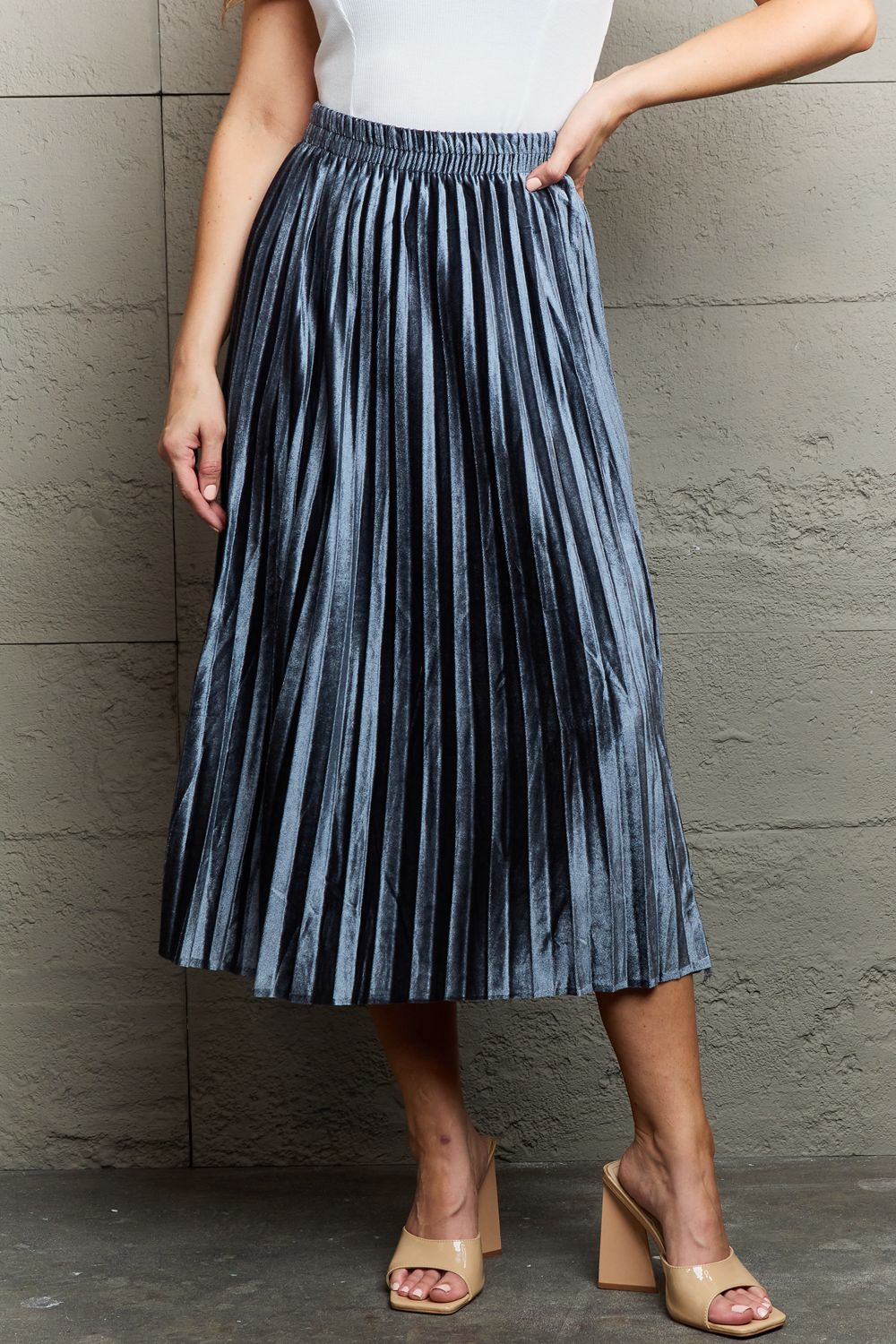 A person in a white top and the Ninexis Accordion Pleated Flowy Midi Skirt stands against a textured wall, wearing beige heeled sandals.