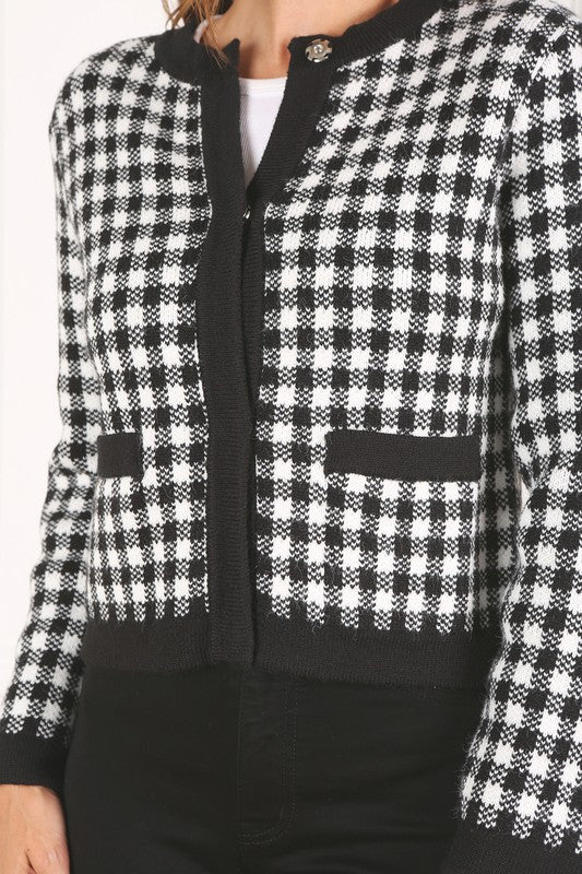 A woman dressed in the Black Check Knitted Jacket, featuring snap buttons and functional pockets, complemented by black pants, stands against a white background.