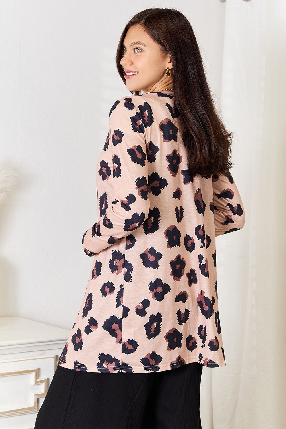 A woman wearing the Double Take Printed Button Front Longline Cardigan, featuring a pink floral pattern, over a black dress, stands indoors near a white vase with dried plants.