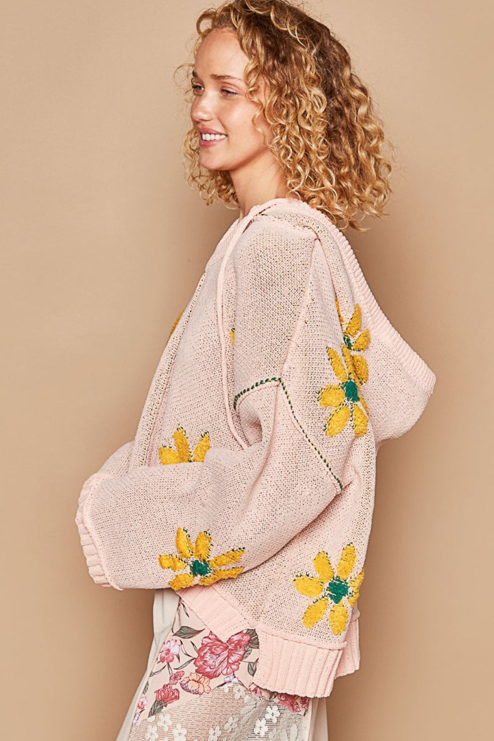 A person with curly blonde hair wears a stylish POL Floral Pattern Hooded High-Low Sweater featuring yellow flower designs and light-colored floral pants. The background is light beige.