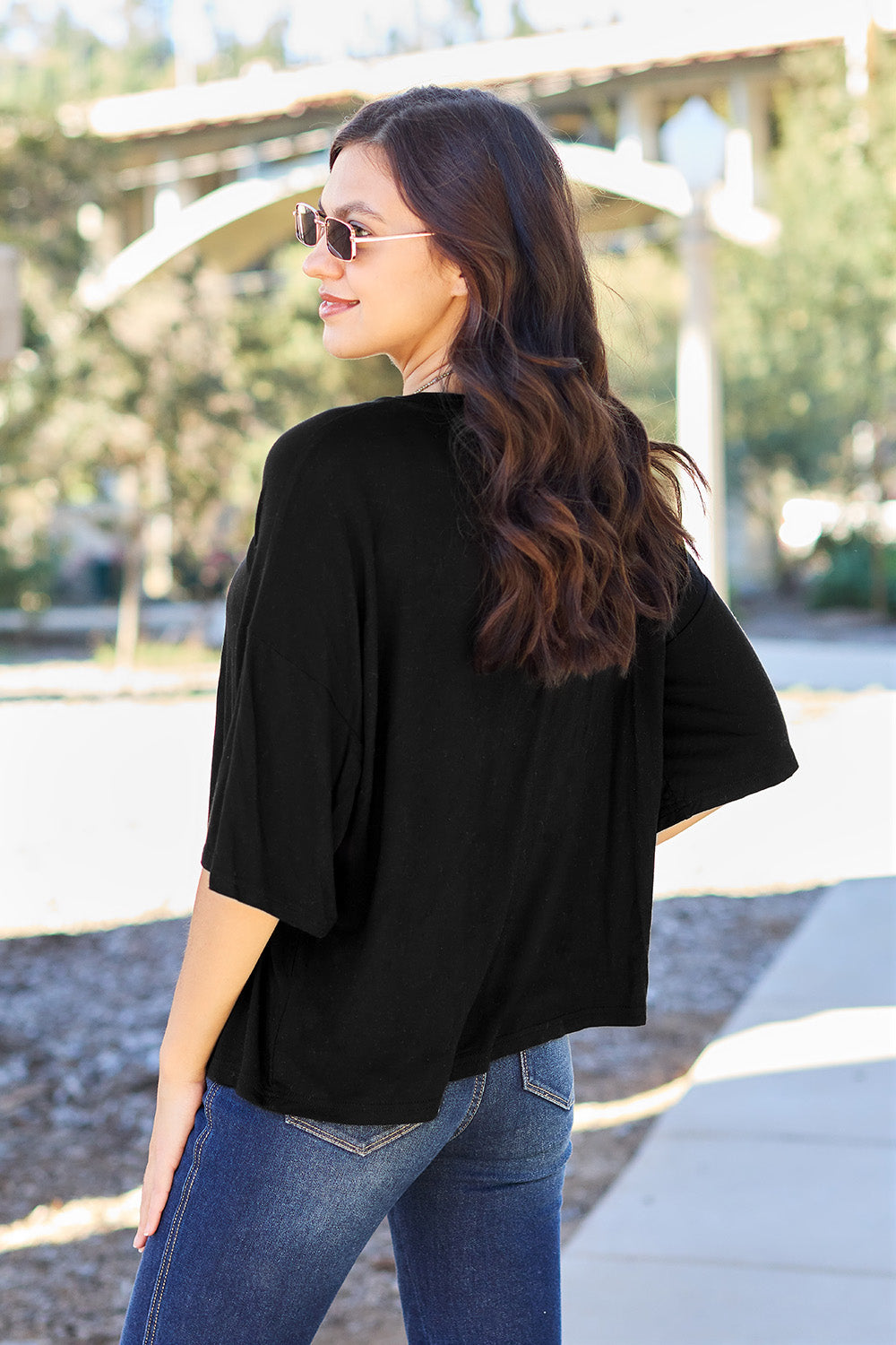 A person with long dark hair and a bright smile stands outdoors, showcasing their basic style in a loose, slightly stretchy *Basic Bae Full Size Round Neck Drop Shoulder T-Shirt* in pink and blue jeans, with one hand on their hip.