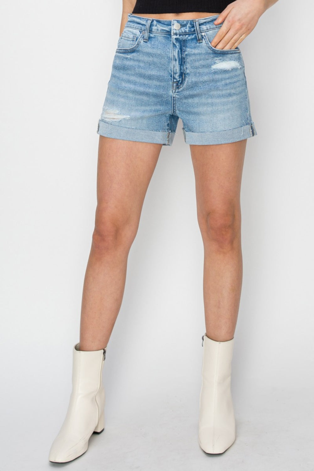 A person wearing RISEN distressed mid-rise waist denim shorts in light blue paired with white ankle boots and a black top, standing against a plain background—a staple for any summer wardrobe.