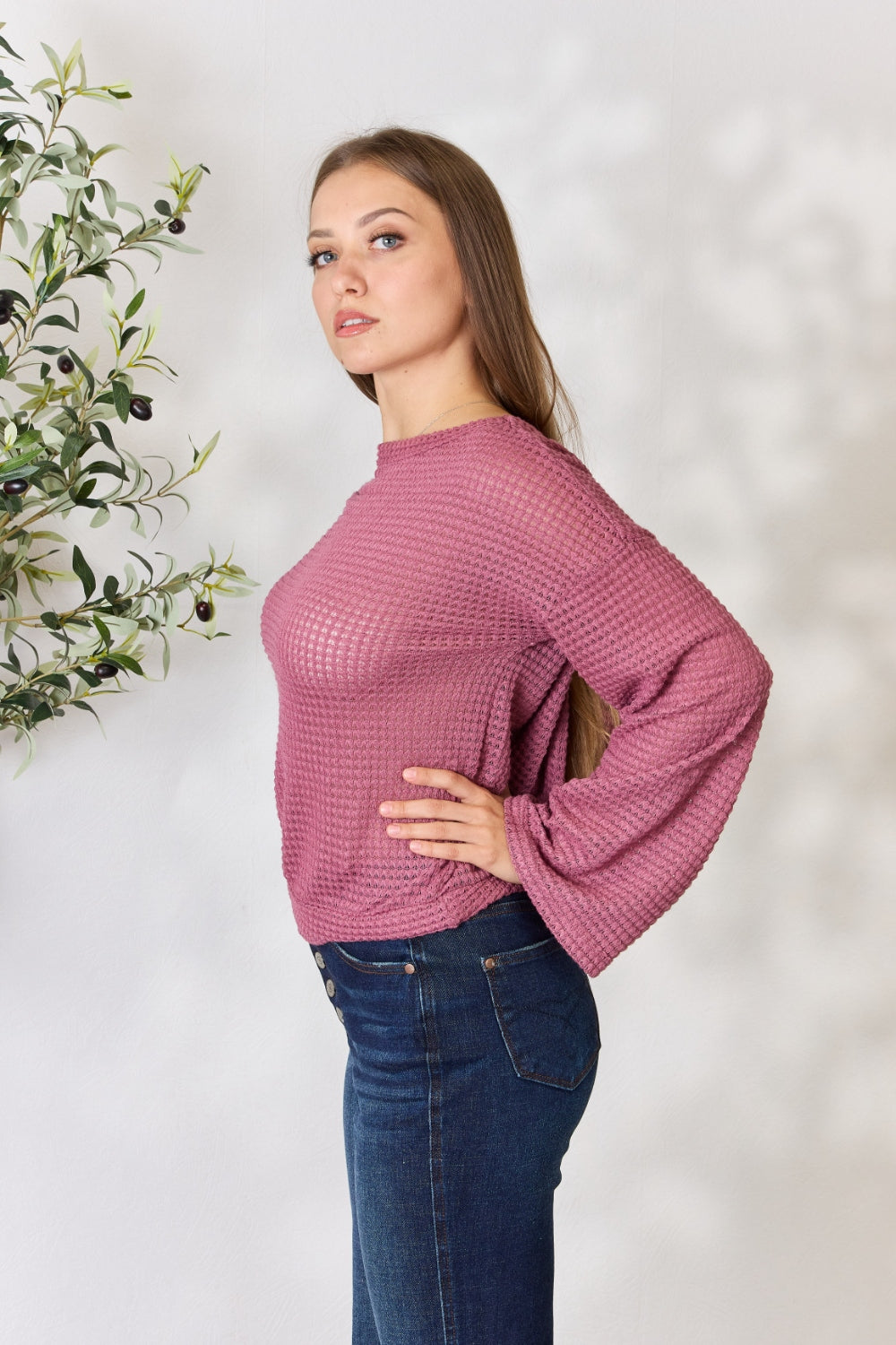 A person wearing a Culture Code Full Size Waffle-Knit Round Neck Long Sleeve Blouse in pink and jeans stands in front of a light background, complemented by a leafy plant on the left.