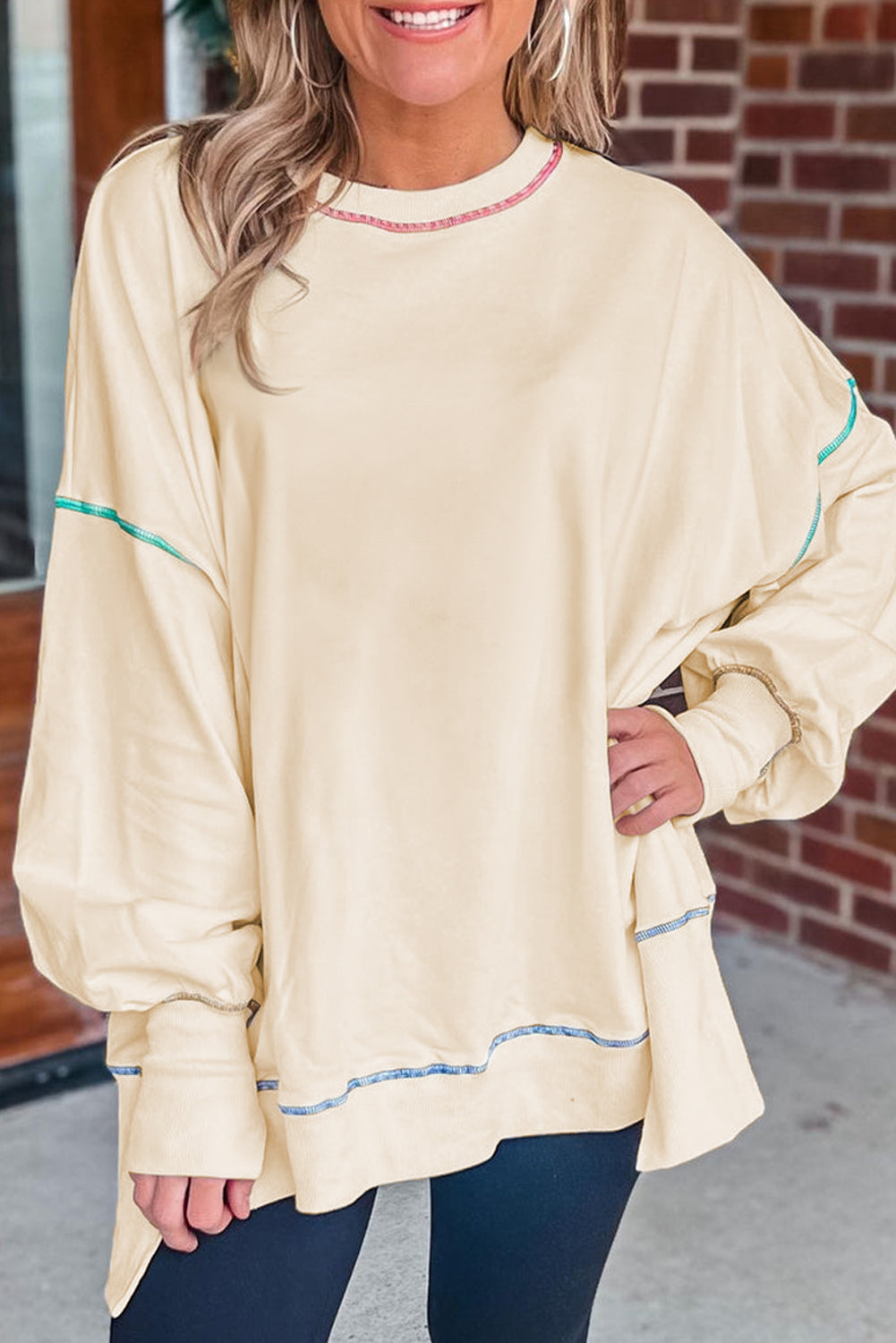 A woman with blonde hair is facing away, wearing a Contrast Stitching Split Baggy Sweatshirt. She stands in a room adorned with plants and wooden furniture in the background.