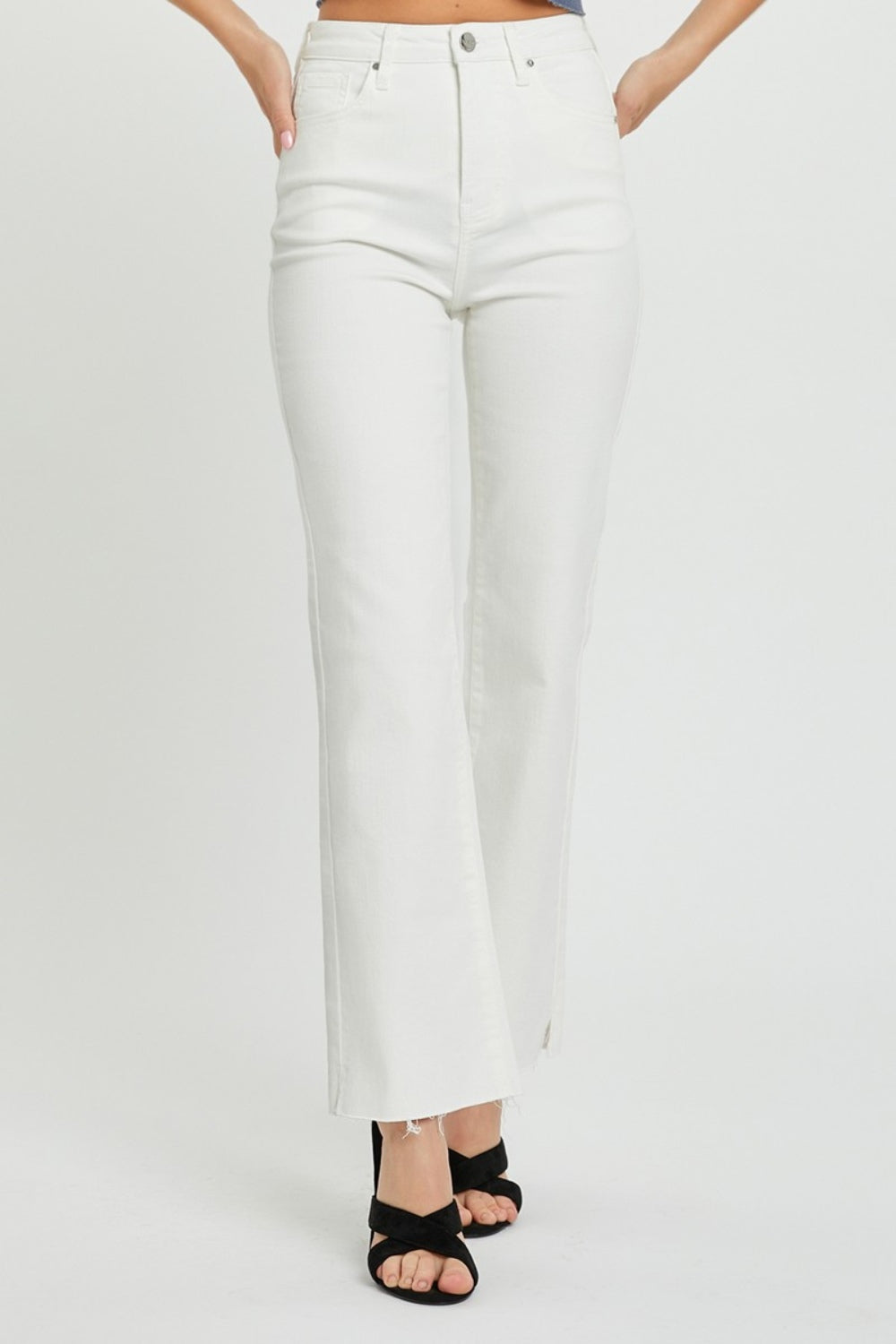 A person wearing RISEN Full Size High Rise Tummy Control Straight Jeans in white with a raw hem and black open-toe heels is standing against a plain background.