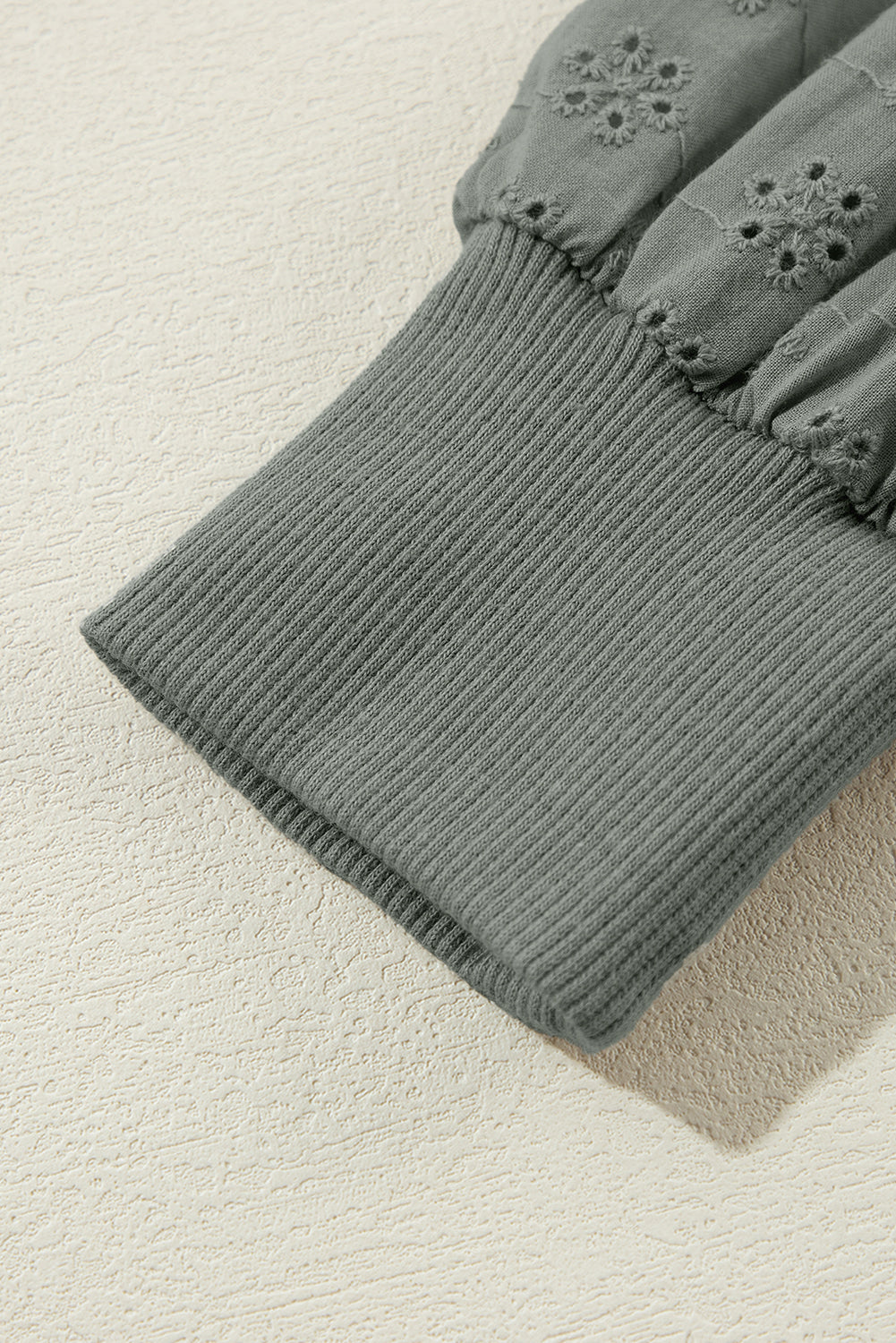 A woman with wavy blonde hair is seen from behind, wearing a grayish-green round neck sweatshirt with patchwork sleeves and blue jeans. She carries a brown shoulder bag against a backdrop of woven wall decorations.