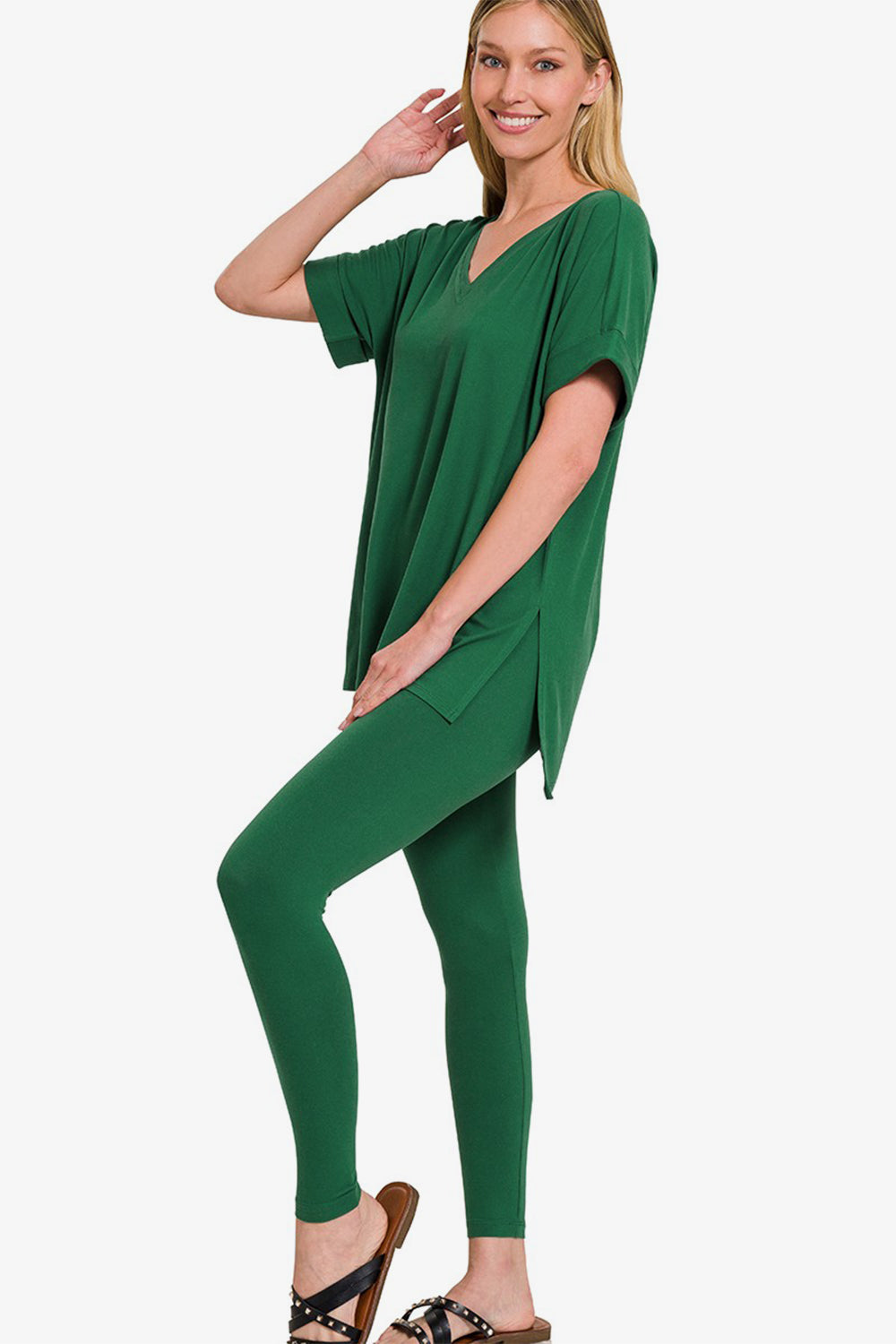 A person wearing the Zenana V-Neck Rolled Short Sleeve T-Shirt and Leggings Lounge Set in green, standing and smiling with one hand touching the side of their head, stylishly matched with sandals.