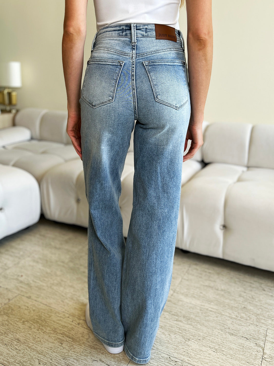 A person wearing Judy Blue Full Size High Waist Wide Leg Jeans and white shoes stands with hands in pockets. They are in front of a white couch and a green plant.