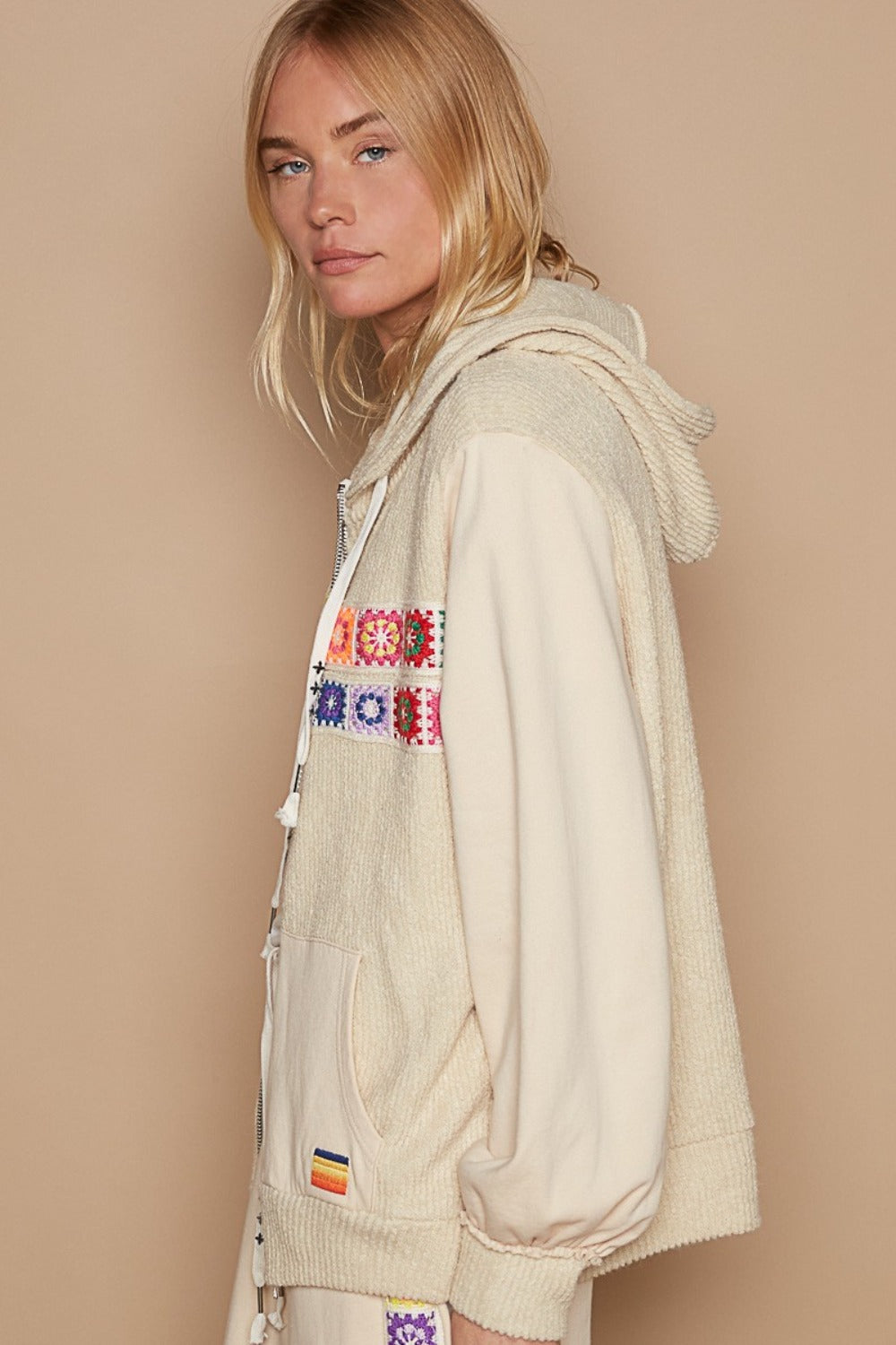 A person with curly hair stands in front of potted cactus plants, wearing the POL Contrast Square Patch Pattern Hooded Zipper Jacket. They pair it with beige pants and a green top underneath, creating a stunning look that showcases fashionable outerwear.