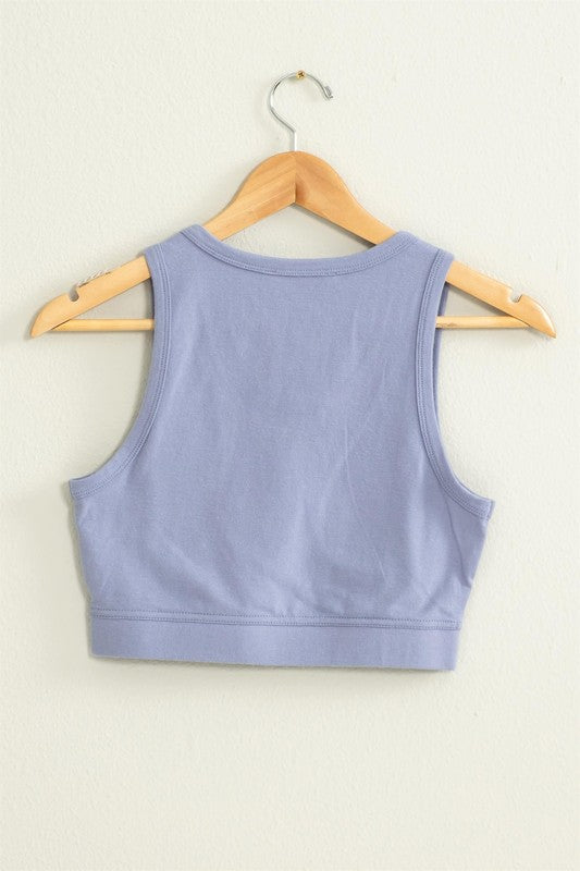 The "All I Need Cropped Tank Top," featuring a light blue hue and a fitted silhouette, hangs on a wooden hanger against a white wall.