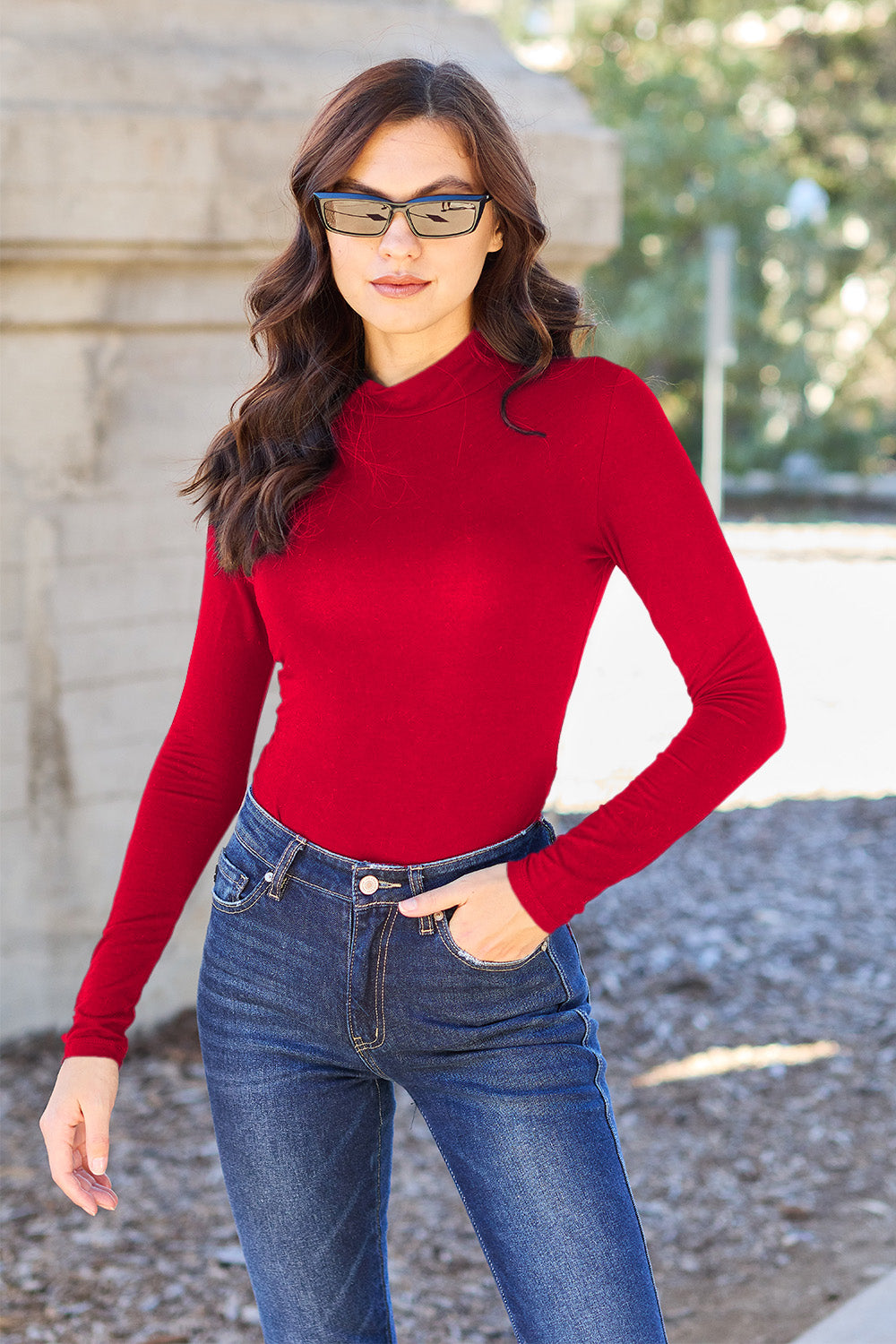 A woman with long, wavy hair stands in front of a light-colored curtain, wearing the Basic Bae Full Size Mock Neck Long Sleeve Bodysuit that offers a basic style and moderate stretch.