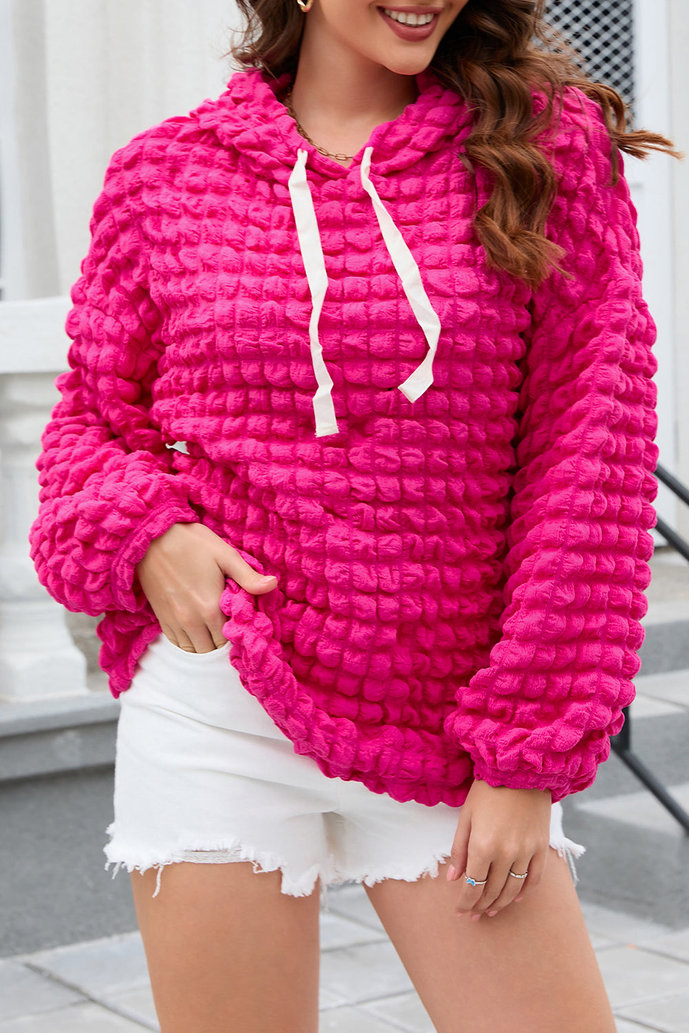 A person stands with their back to the camera, donning the Rose Bubble Textured Waffle Hoodie in a vibrant pink hue. They are in a room with yellow floral wallpaper, and a corner of a wooden chair is visible.