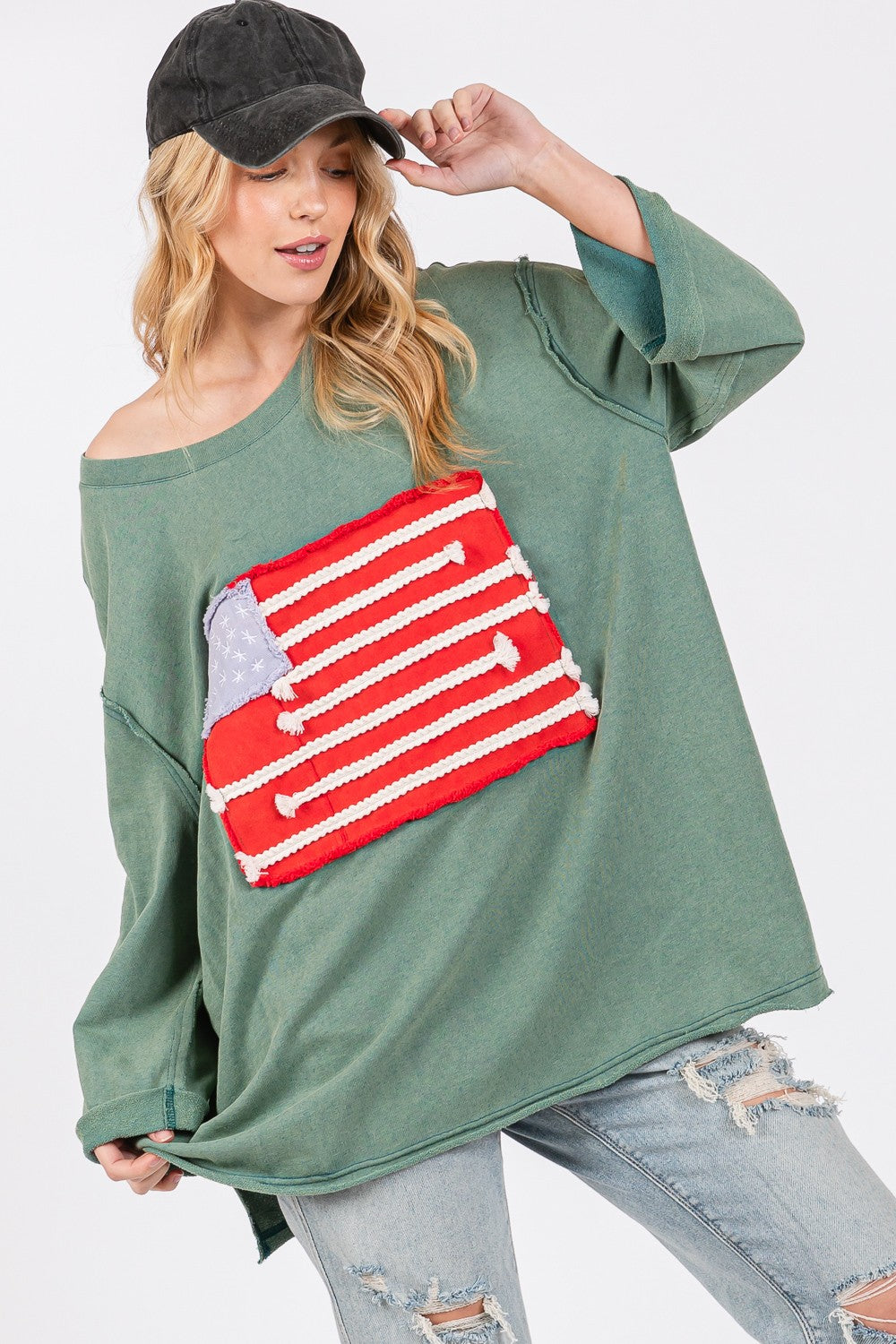 A person exudes casual charm in the SAGE + FIG Full Size American Flag Patch Drop Shoulder T-Shirt, paired with ripped jeans, confidently posing with hands in hair against a plain background.