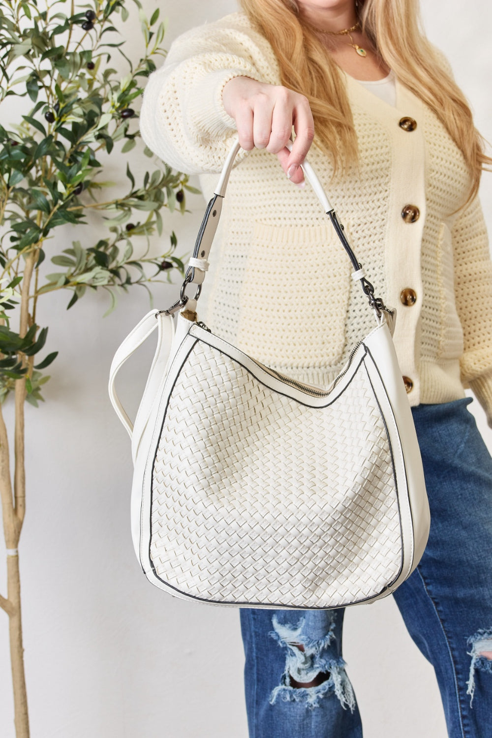 A person carries the SHOMICO Weaved Vegan Leather Handbag in a stylish brown hue, featuring a sleek crossbody strap, expertly paired with a cream sweater and jeans.