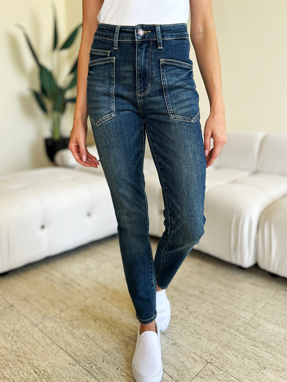 A person wearing Judy Blue Full Size High Waist Skinny Jeans made of stretchy denim fabric stands in a room with light-colored furniture and wooden flooring, paired perfectly with white slip-on shoes.