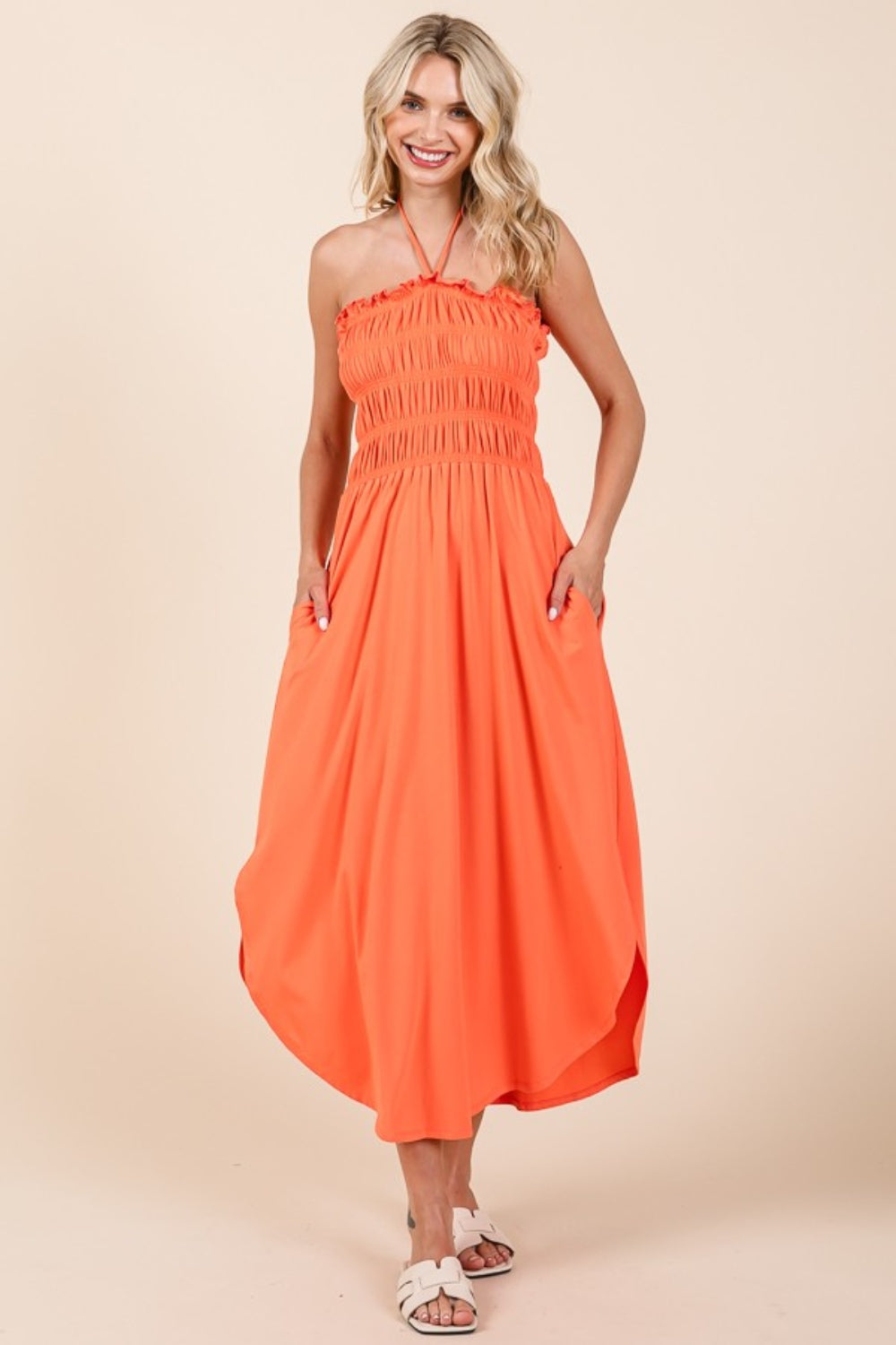 A woman wearing the Culture Code Tie Back Shirring Dress with Pockets, a versatile wardrobe staple in orange, stands against a plain background, smiling with her hands comfortably resting in the pockets.