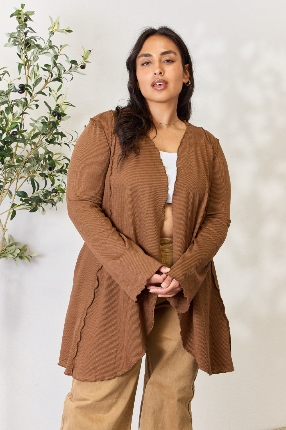 A woman stands in front of a light-colored wall and a small green plant, wearing the Culture Code Full Size Open Front Long Sleeve Cardigan with a merrow edge finish over a white top and beige pants.