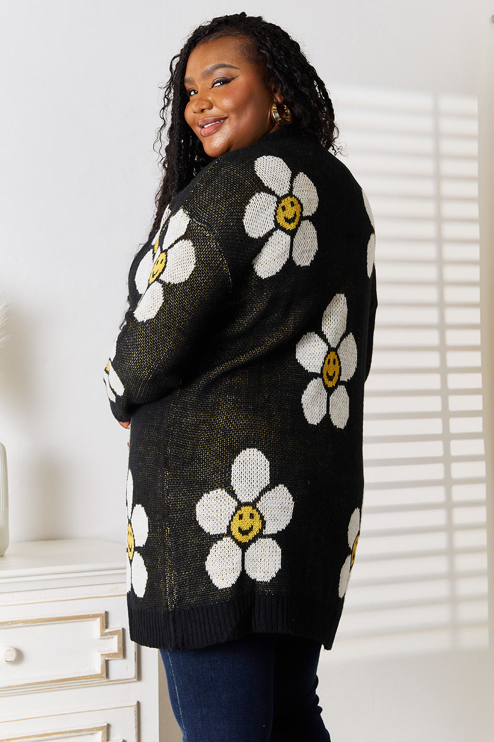 A woman stands indoors wearing the Perfee Floral Button Down Longline Cardigan, which features stylish black fabric adorned with white and yellow smiley face flowers. She pairs it with a white top and blue jeans, looking off to the side while smiling.