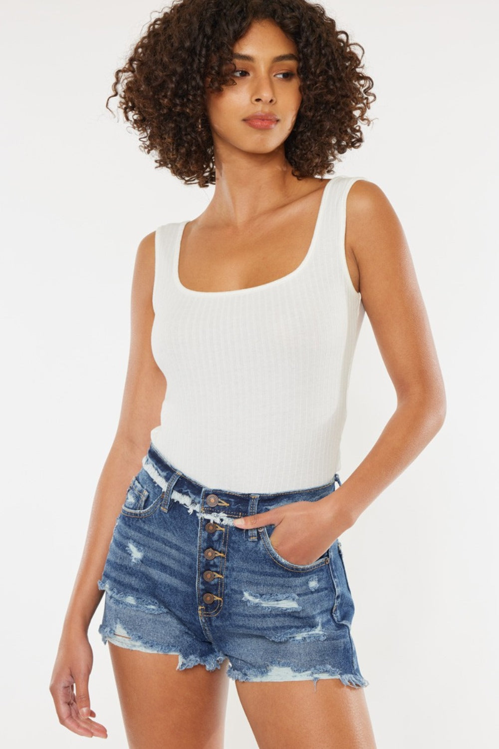 A woman with curly hair wearing a white sleeveless top and Kancan Distressed Button Fly Denim Shorts stands against a plain background, perfect for a stylish summer wardrobe.