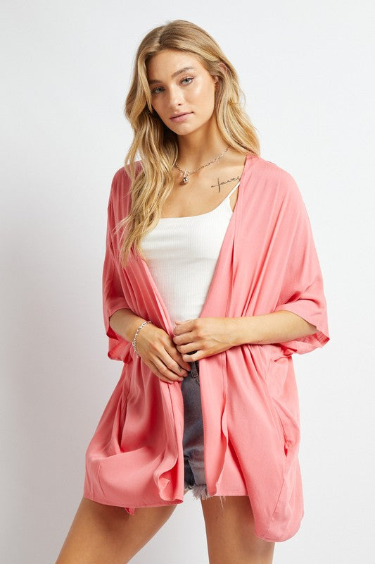 A woman poses against a simple backdrop, gracefully wearing a wardrobe essential: the Favorite Solid Kimono Cardigan in pink. Complementing her white tank top and denim shorts, this casual layer enhances her relaxed style with an elegant touch.