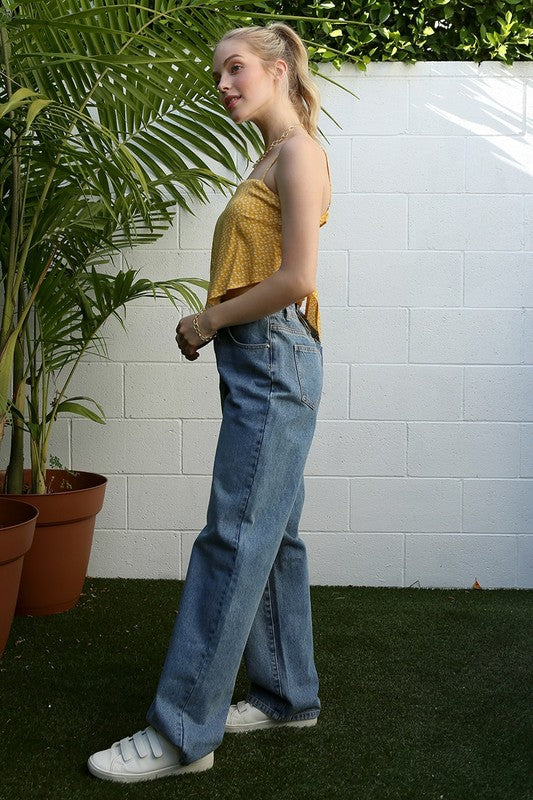 A person wearing an SL yellow floral tank top and jeans stands by a palm plant against a white wall.