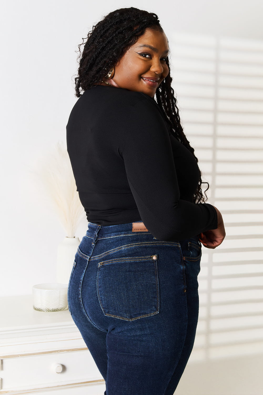 A woman with long hair is wearing a modern, stylish wardrobe featuring the Basic Bae Asymmetrical Neck Long Sleeve Top and jeans. She stands indoors in front of a white wall with shadow patterns.