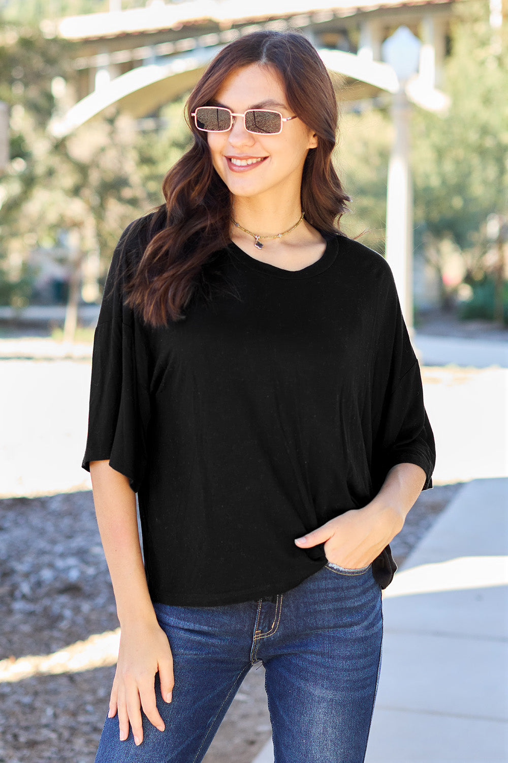 A person with long dark hair and a bright smile stands outdoors, showcasing their basic style in a loose, slightly stretchy *Basic Bae Full Size Round Neck Drop Shoulder T-Shirt* in pink and blue jeans, with one hand on their hip.