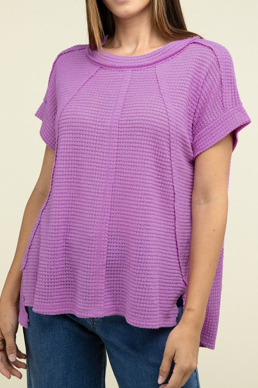 A woman with long brown hair is wearing a purple Brushed Waffle Exposed-Seam Short Sleeve Top and blue jeans, standing against a plain beige background. This casual wardrobe choice, with its short sleeves and side slits, is perfect for everyday wear.