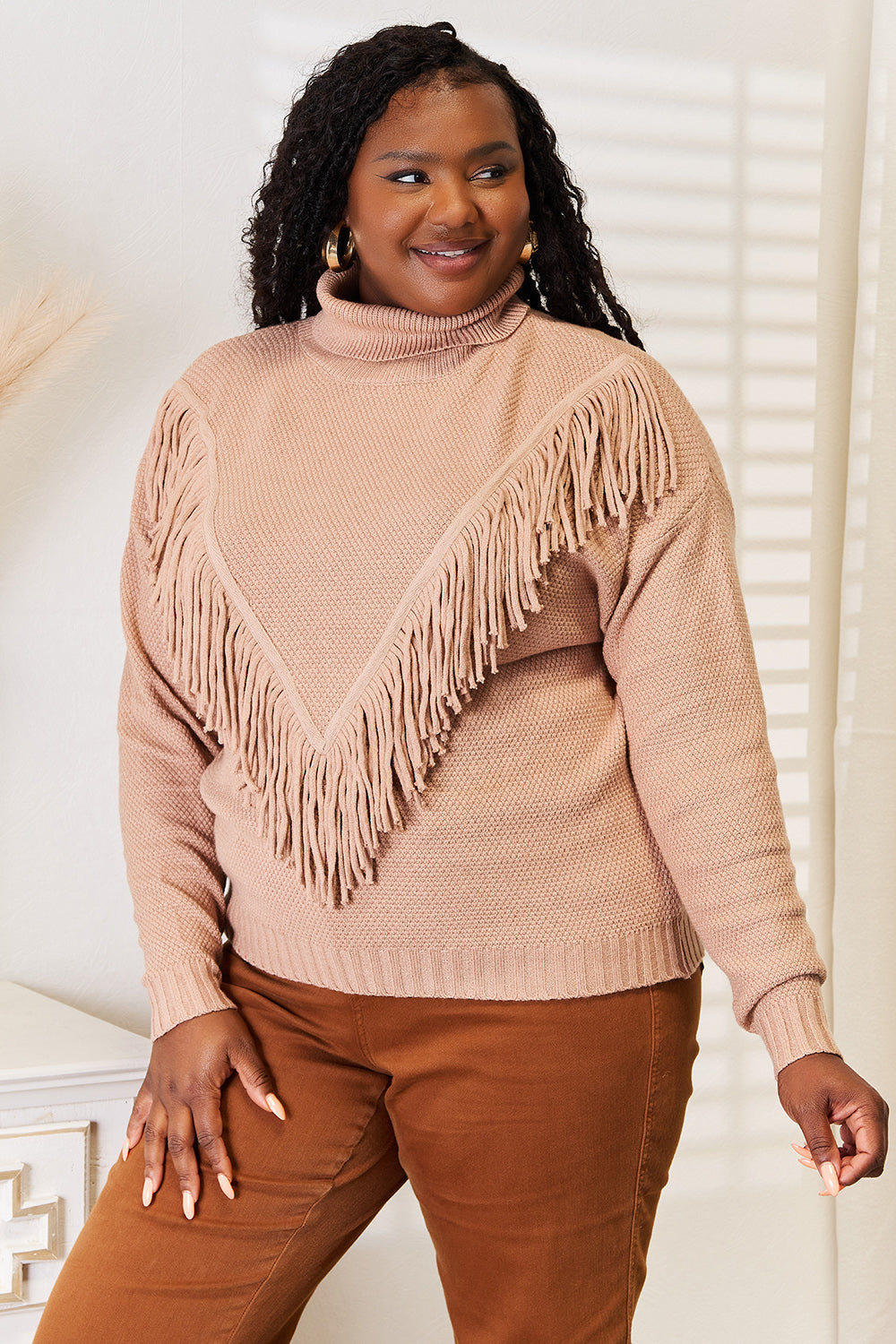 A woman wearing the Woven Right Turtleneck Fringe Front Long Sleeve Sweater, her winter essential, pairs it effortlessly with brown pants. She stands indoors with a neutral expression, embodying cozy sophistication.