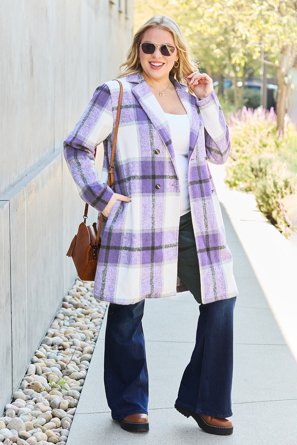 A woman stands on a sidewalk wearing the Double Take Full Size Plaid Button Up Lapel Collar Coat in pink and white over a white top and blue jeans, with her right hand lightly touching her hair. The polyester fabric of the coat makes it durable and perfect for an easy machine wash cold.