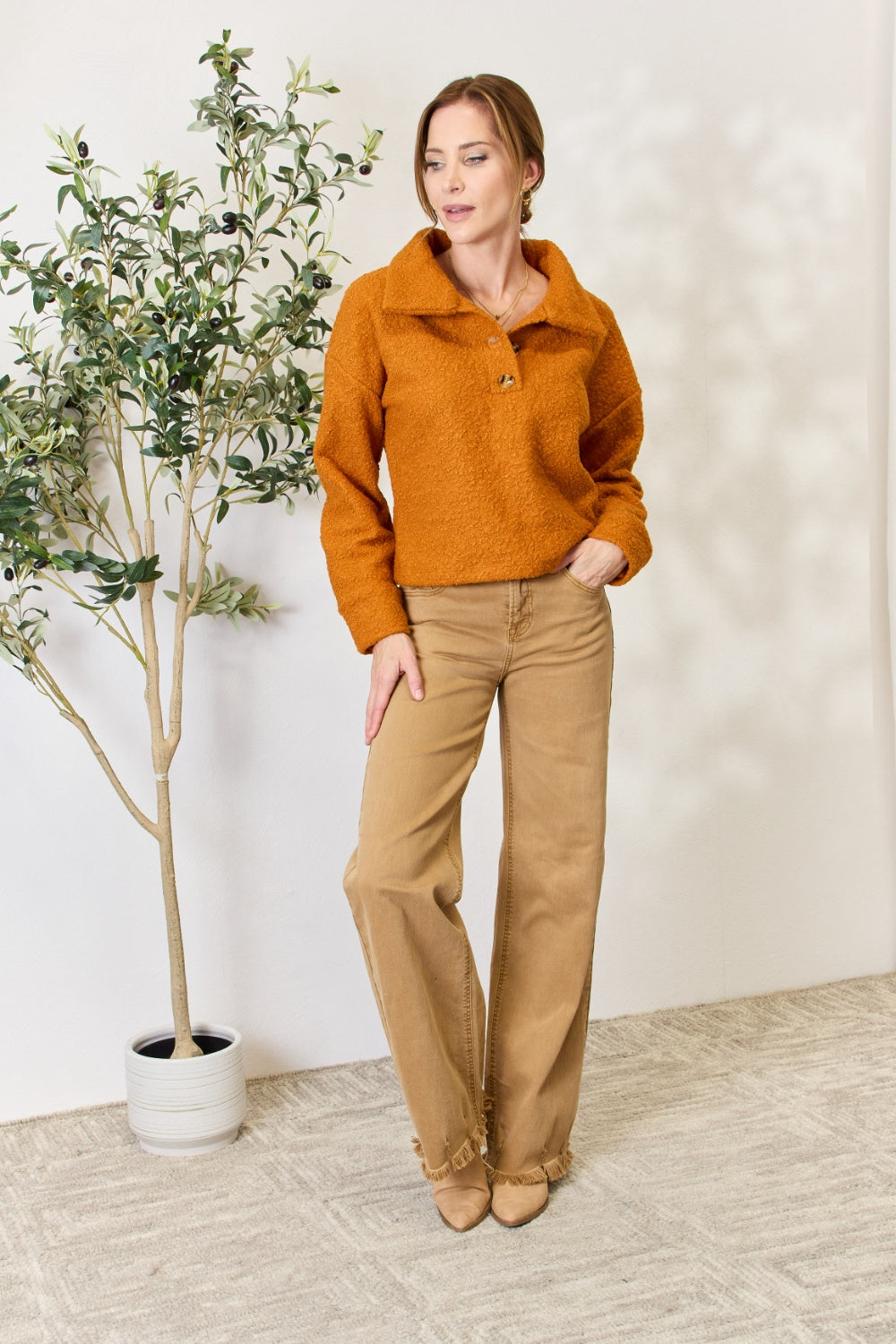 A person wears a brown textured Culture Code Full Size Half Button Turtleneck Sweatshirt made from breathable fabrics and tan pants, standing next to a leafy plant against a light background.