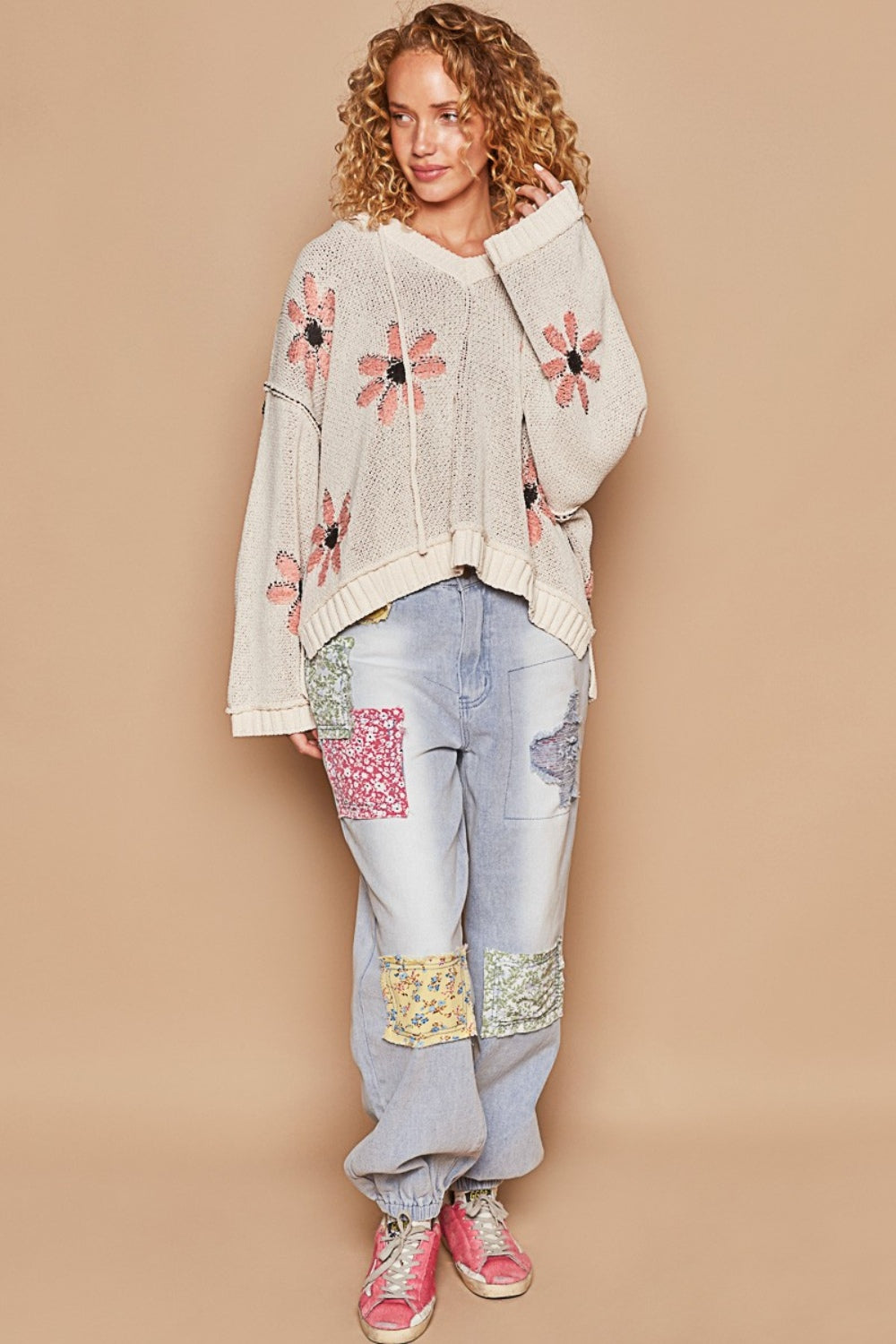 A person with curly blonde hair is sitting on a wooden stool, smiling, and wearing the POL Floral Pattern Hooded High-Low Sweater paired with light blue jeans with patches. The background is beige.