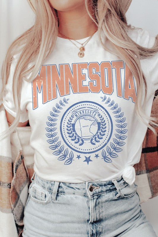 A woman sporting a 100% cotton MINNESOTA Graphic Tee with "Minnesota" emblazoned in bold orange letters and a decorative state emblem underneath, complemented by high-waisted denim shorts and a plaid shirt casually draped over her shoulders. The tee offers unisex sizing for an extra comfortable fit.