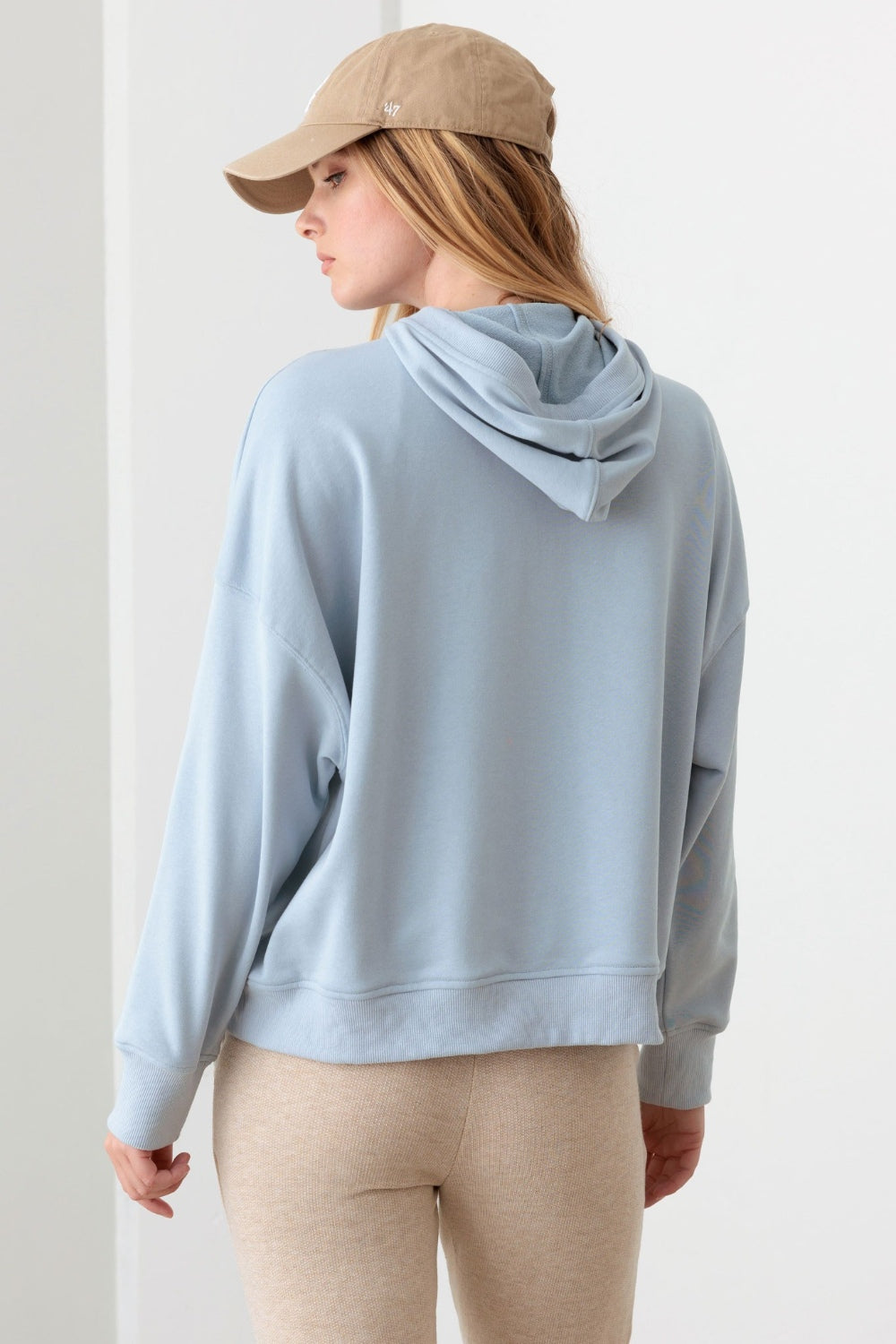 In a neutral-colored room, a person is wearing the Le Lis Half Zip Drawstring Mock Neck Hoodie in light blue along with a beige cap.