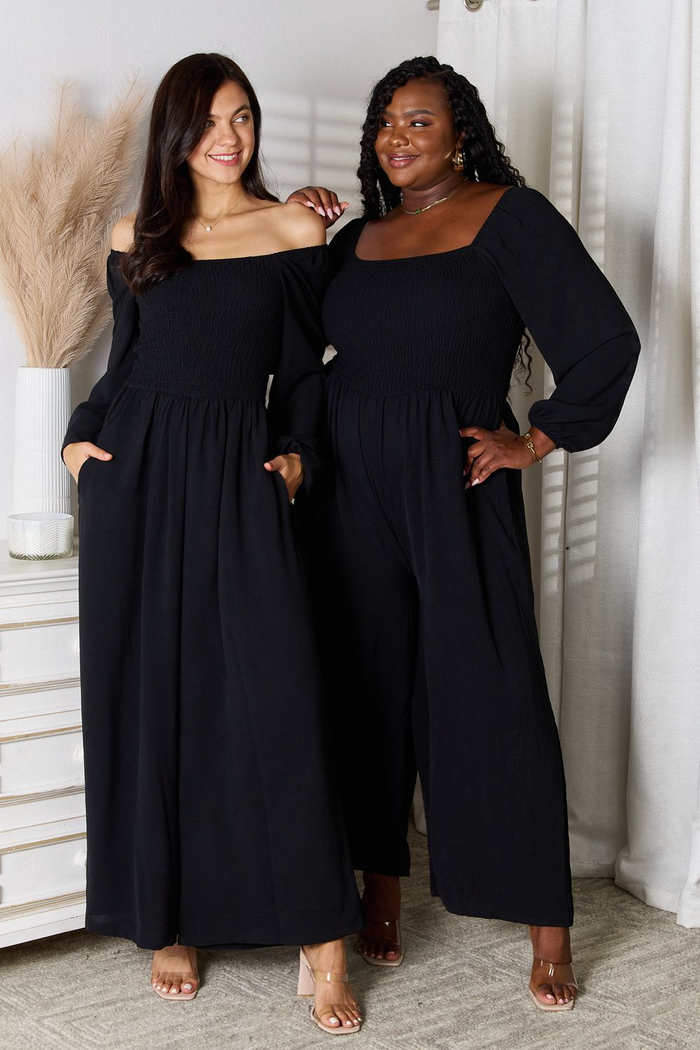 Two women in Double Take Square Neck Jumpsuits with pockets stand side by side in a room decorated with white accents and pampas grass in a vase. Both are smiling, have their hands in their pockets, and are wearing high heels.