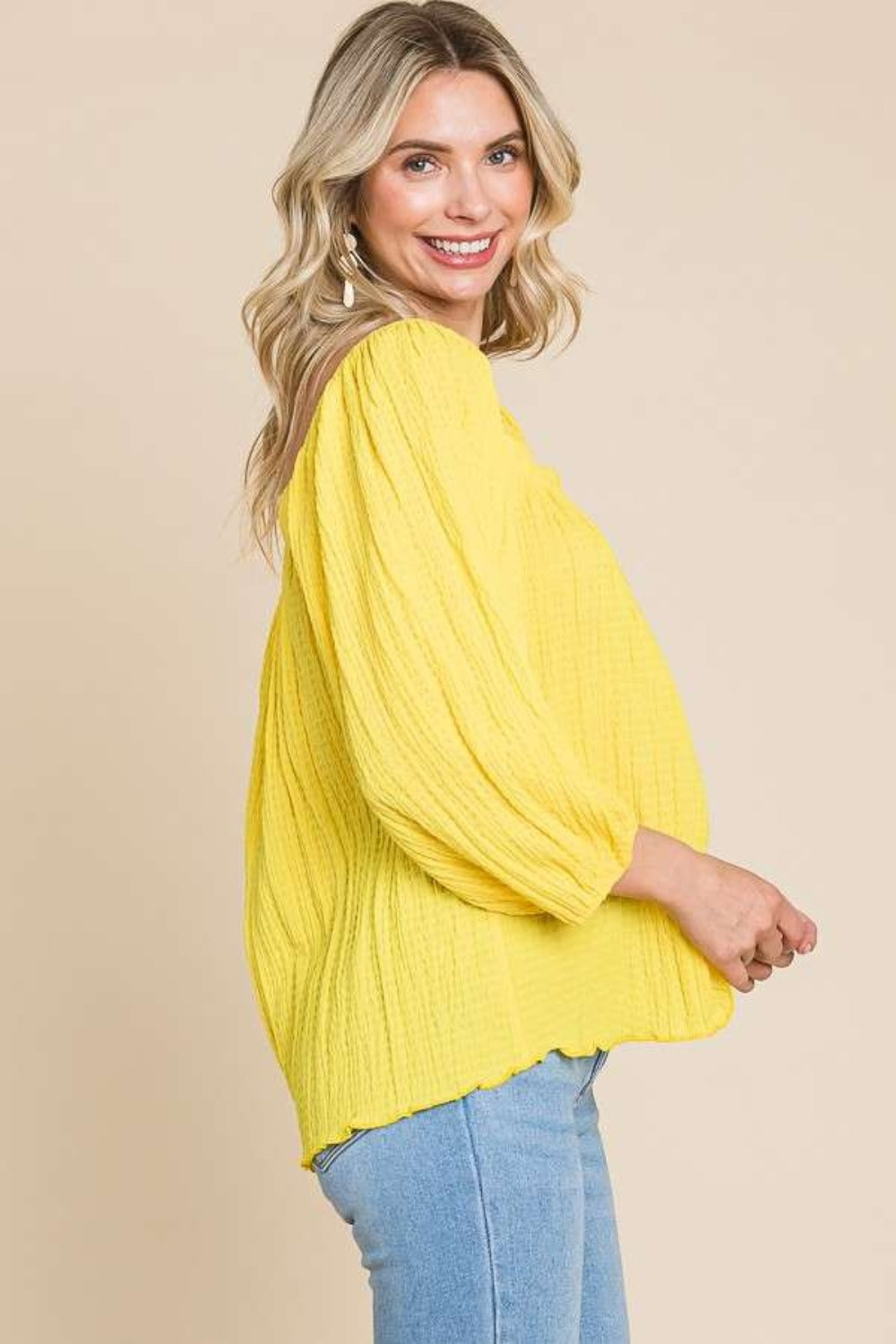 A woman wearing a yellow Culture Code Texture Square Neck Puff Sleeve Top and blue jeans stands against a beige background.