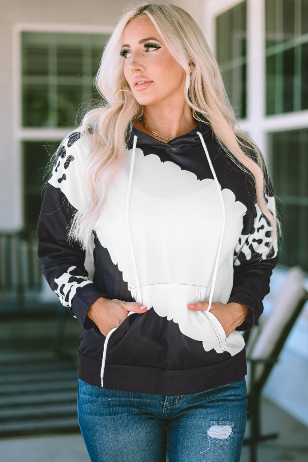 A person with long blonde hair is seen from the back wearing a Black Cow Tie Dye Print Pocketed Drawstring Pullover Hoodie and blue jeans. They are standing in an indoor space with a window and furniture in the background.