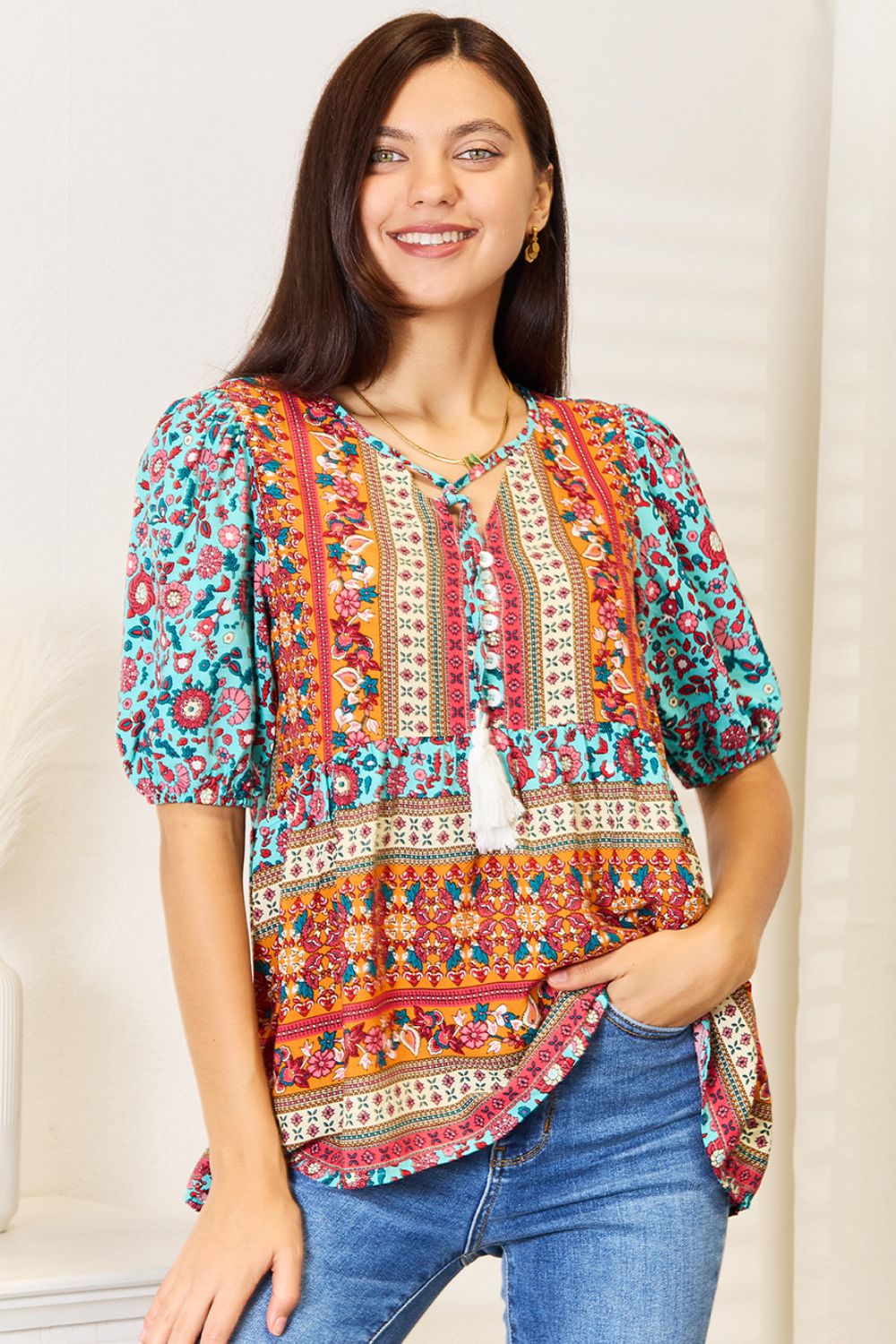 A woman wearing the Devine Bohemian Tassel Tie Puff Sleeve Babydoll Blouse, adorned with a mix of vibrant floral designs, smiles in her jeans while standing indoors against a light background.