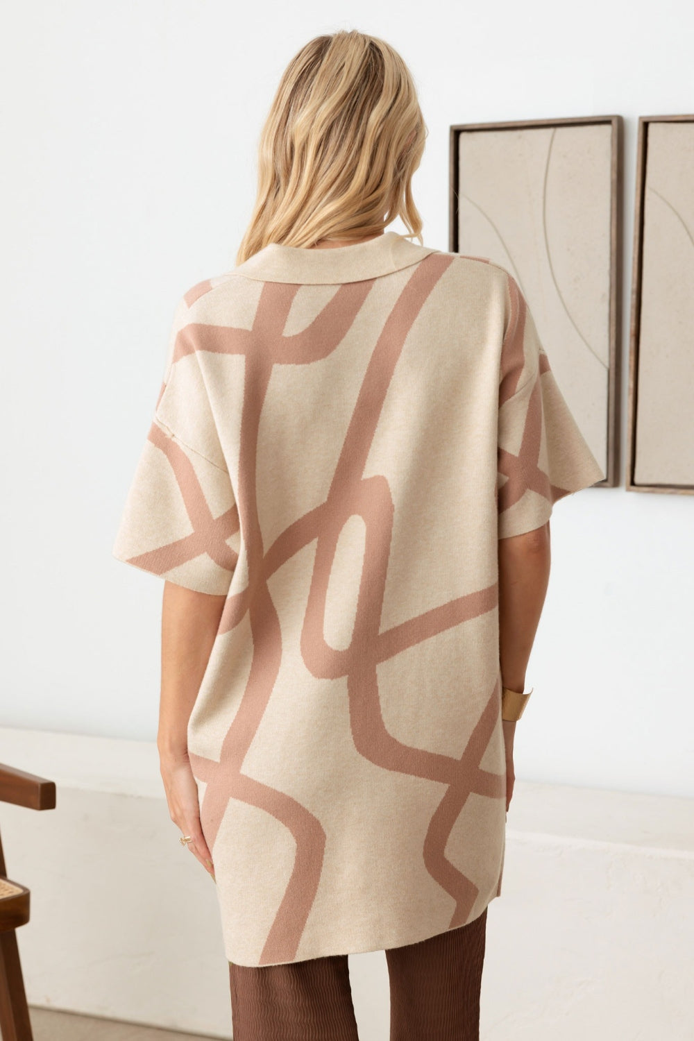 A woman with long blond hair wearing the Le Lis Abstract Collared Button Down Sweater Dress stands in a minimalist room with a wooden chair and ceramic vase—her versatile piece adds an abstract touch to the setting.