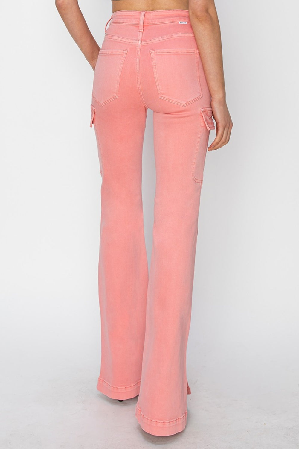 A person wearing RISEN Full Size High Rise Side Slit Cargo Bootcut Jeans, featuring a pink flared design with front pockets, stands against a plain background.