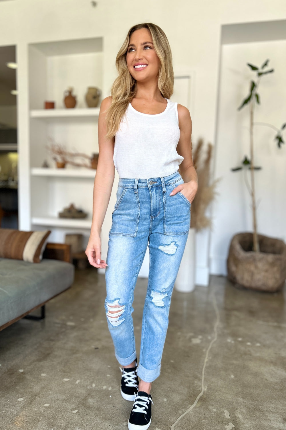 Two women are standing side by side, both clad in black and white tank tops and Judy Blue Full Size Distressed Straight Jeans with Patch Pockets, smiling at the camera in a modern living room.