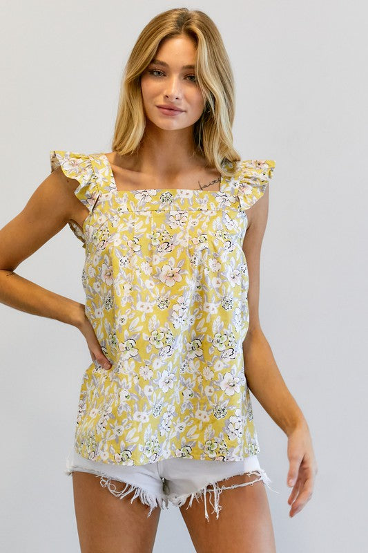 A woman in a vibrant summer wardrobe stands against a plain background, wearing the Floral Printed Ruffle Sleeveless Top paired with white shorts, with one hand casually tucked in her pocket.