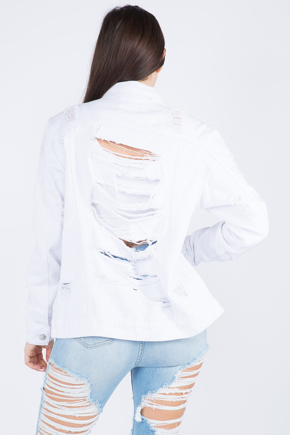 A woman wearing an American Bazi Distressed Back Button Down Denim Jacket over a white crop top and distressed blue jeans stands with her left hand holding the jacket collar.